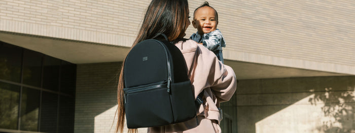 Mom wearing a black backpack and holding a baby outside.
